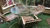 Overview of round glass top outdoor table and four floral cushioned chairs on a porch.
