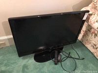 Front view of Samsung TV turned off on black stand with two remotes and power cord on green carpet next to floral couch.