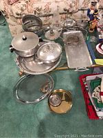 Photo showing various hammered aluminum cookware pieces including covered casseroles, round trays, rectangular tray, and other kitchenware items on glass table.