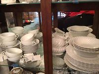 Photo of multiple white china cups, saucers and bowls mostly stacked inside a glass cabinet. The cups have subtle floral patterns showing the Kaiser brand style.
