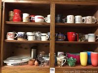 Wood cabinet shelf holding 20+ assorted coffee cups and mugs, including colorful, white, and black mugs. Pyrex baking dish with lid on lower shelf with small pottery and glasses nearby.