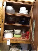 Wood kitchen cabinet open showing multiple glass and Pyrex casserole dishes in clear, amber, and blue along with white and green baking dishes, glass measuring cup, and red-handled utensil set.