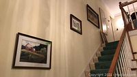 View of five framed artworks hanging along stairwell wall, various sizes and subjects.