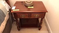 Front and top view of the wooden side table with two drawers and brass handles, along with a lamp base on top (lamp not included). Table next to a bed.