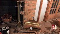 Brass fireplace tools set standing next to a brick fireplace, including poker, shovel, broom with animal-head handles, and a brass mesh ball hanging from the stand. Also visible is a curved brass wood basket with handle on the floor.