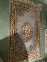 Photo of a rectangular rug featuring a central oval floral medallion, light blue background, ornate floral borders, and beige fringe on one end.
