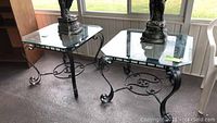 Pair of square wrought iron end tables with glass tops on porch floor, showing overall design and size.