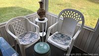 Two white plastic garden chairs with floral cushions, ceramic round plant stand, brass and epoxy urn on a white pedestal plant stand. Screened porch view with lawn outside.