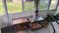 Two wood end tables, one with glass insert and wicker shelf, the other on rollers, placed on porch floor with large ceramic vase on glass-top table