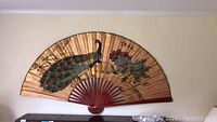 Front view of the large oriental style paper fan hanging on the wall, showing the peacock and floral painted design between wooden slats.
