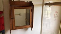 Front view of wooden medicine cabinet mounted on wall showing framed mirrored door with wooden knob and decorative top edge.