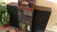 Front view of the matched pair of Curtis Mathis cabinet speakers positioned beside a wooden storage unit and radio on top.