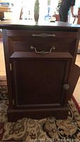 Front view of one composite wood side table showing cabinet door with metal hinge and knob, drawer with decorative metal handle, and part of the base and top surface.