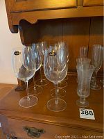 Photo of multiple clear glass wine glasses with long stems, some with Accademia Italy stickers, and parfait glasses grouped together on a wood surface.