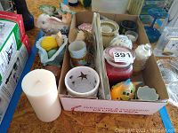 Photo showing a box full of assorted candles, candle holders, and ceramic decorative items including a ceramic bunny and colorful ceramics.