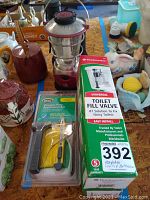 Photo of items on a table including a red and silver GE propane lantern, boxed Fluidmaster toilet fill valve kit, sealed Gila window film application kit, and assorted decorative ceramic pieces including pigs and ducks.