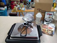 Gotham Steel indoor grill with solid and slotted grill plates, GT Xpress 101 cooker with box and accessories, user manual, and measuring cup on table.