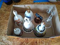 Eight collectible decorative bells in a cardboard box, made from lead crystal and ceramic with floral designs.