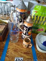 Photo showing two German beer steins with lids and a clear glass beside them on a table with various household items.