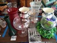 Overview of assorted glass and plastic vases with some containing faux greenery and other fillers.