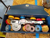 Dymo-Mite motion metal label maker with various colored tape rolls in original blue carrying case on a wood surface with some wear.