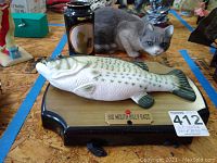 Big Mouth Billy Bass mounted on wooden plaque with cat figurine statue behind it.