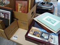 Various framed pictures and prints packed or stacked, several wooden picture frames. A green tinted print visible on top, cardboard boxes contain other framed prints.