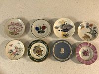 Top view of eight mini decorative plates arranged in two rows on a white surface, showcasing various designs including floral, landscape, and classical motifs.