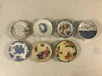 Top view of seven mini antique china plates arranged in two rows with various colorful floral, bird, fruit, and scenic patterns.