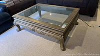 Square powder coated metal frame coffee table with bevelled glass inset top viewed from front corner showing ornate carved leg detail.