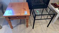 Photo showing two occasional tables side by side: pine wood table with drawer and metal frame table with glass top on carpeted floor.