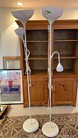 Front view showing two white metal floor lamps with round base, tall poles, and dual lights, one bowl-shaped upward and one cone-shaped adjustable side light.
