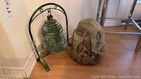 Pair photo showing the clay bell with metal stand and carved stone sculpture side by side on a wood floor by a wall corner.