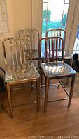 Four chrome metal bar stools grouped together on wood flooring near glass doors.