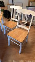 Two painted wood chairs with natural wood seats, showing wear including scuffs and chips. Chairs have vertical spindles and curved top backs, painted white.