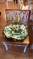 Wooden artisan tray with yellow bird figurine on top and a rectangular metal tray with olive oil bottle illustrations leaning against a chair back