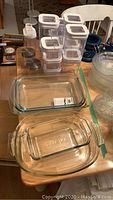 View showing 5 glass casseroles, some with lids, and 13 piece Fontal plastic storage containers with white lids on wooden table.