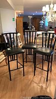 Front view of all three black metal bar stools with vinyl seats and vertical slat backs, standing on hardwood floor.