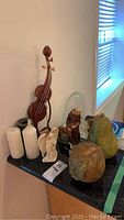 Carved wood violin sculpture with broken neck, angel figurine, monk figurine, three white cylindrical candles, and clay fruit sculptures displayed on granite counter near window.