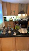 Photo of metal candle centrepiece with 3 hanging glass votive holders, metal candlestick holders, silver goblets, and ornate silver dish on a kitchen island.