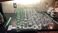 Wide view of approximately 70 assorted stemware glasses including clear and green-tinted glassware arranged on a dark countertop.