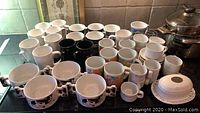 Photo of 22 assorted mugs, 4 soup cups, butter dish and espresso set arranged on kitchen counter.