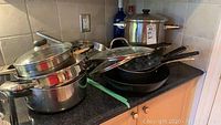 Photo showing stacked pots and pans on kitchen counter, some with glass lids and long handles.