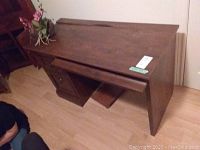Front-left angle view of the faux wood desk showing the keyboard tray pulled out and the filing cabinet drawer closed, positioned against a wall with wood flooring.