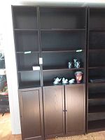 Front view of two tall dark laminate IKEA bookcases with open shelves and lower cabinets with doors. Decor items placed on some shelves.