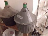 Three large glass wine carboys with green tops inside gray plastic protective baskets with conical lids, positioned on wooden stands.