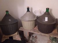 Three glass carboys with green necks and cork stoppers inside ribbed protective plastic baskets (one dark gray, one beige, one brown) placed on wooden shelves in a basement area.