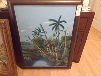 Framed landscape painting showing palm trees, river, and land with cloudy sky, featuring a wooden frame.
