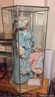 Six-sided antique leaded glass doll display cabinet containing two dolls for scale, featuring hinged lid and tall glass panels.