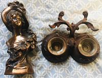 Photo showing the antique cast iron double inkwell with two glass wells and quill pen rests alongside the Victorian Lady spelter clock topper on a patterned fabric surface.